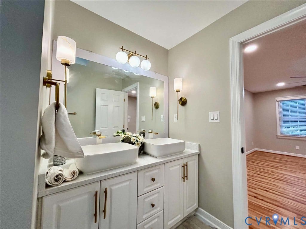 2255 Oak Bay Lane, Henrico, VA 23233 Dual rectangular vessel sinks atop a white vanity with brass hardware, illuminated by a tri-bulb vanity light and matching brass wall sconces, featuring a large wall-mounted mirror