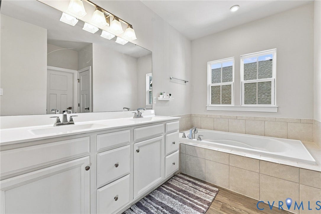 1716 German School Road, Westlake Heights, Richmond, VA 23225 Double vanity with white cabinetry, solid surface countertop, inset soaking tub with tile surround, and large wall-mounted mirror