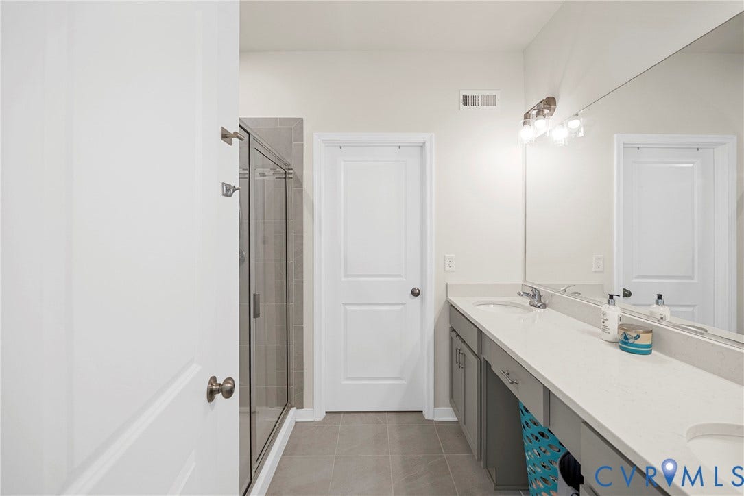 14501 Hancock Towns Drive, Chesterfield, VA 23832 Full-width vanity with a light-toned solid surface countertop and dual undermount sinks, featuring grey cabinetry, a frameless glass-enclosed shower with tile surround, and a wall-to-wall mirror