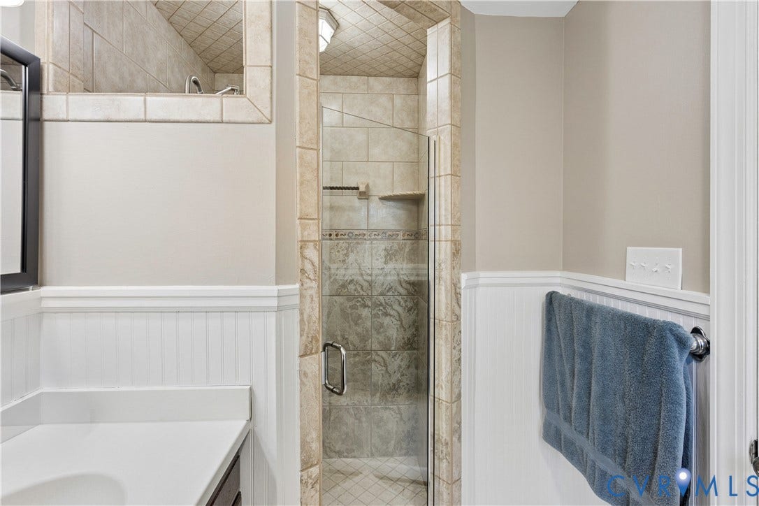327 Huddersfield Drive, Chesterfield, VA 23236 Walk-in shower enclosure featuring tiled surround and stone-finish accent, integrated corner shelving, white beadboard wainscoting, and white vanity countertop
