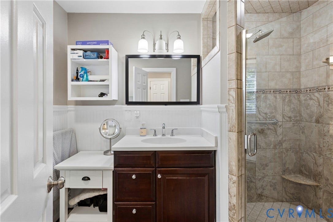 327 Huddersfield Drive, Chesterfield, VA 23236 Bathroom featuring a dark wood vanity with a light-colored countertop, three-light fixture above a framed mirror, built-in white wall shelving, and a tiled shower enclosure with a glass door and integrated seat