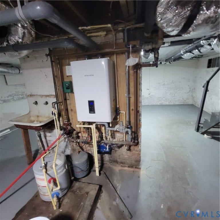 Tankless water heating system featuring a digital display, mounted on a wood backing with exposed utility piping, alongside a wall-mounted utility sink