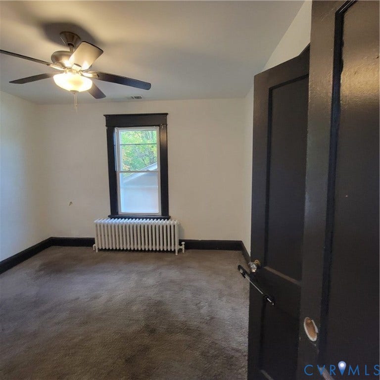 Room featuring carpeted flooring, a ceiling fan with integrated light fixture, dark-trimmed window, and cast-iron radiator