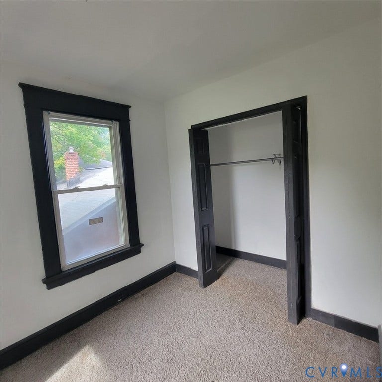 Neutral-toned interior space featuring carpeting, a double-hung window with dark-toned trim, and a built-in closet with folding doors