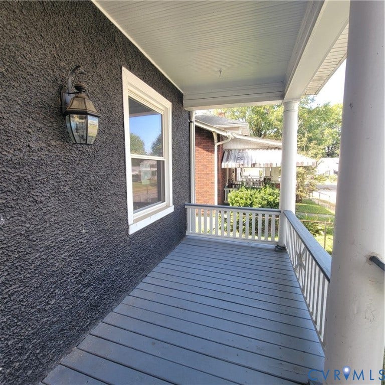 Covered front porch featuring textured dark exterior siding, wood-plank flooring, a decorative column, painted railings, and a wall-mounted carriage-style light fixture