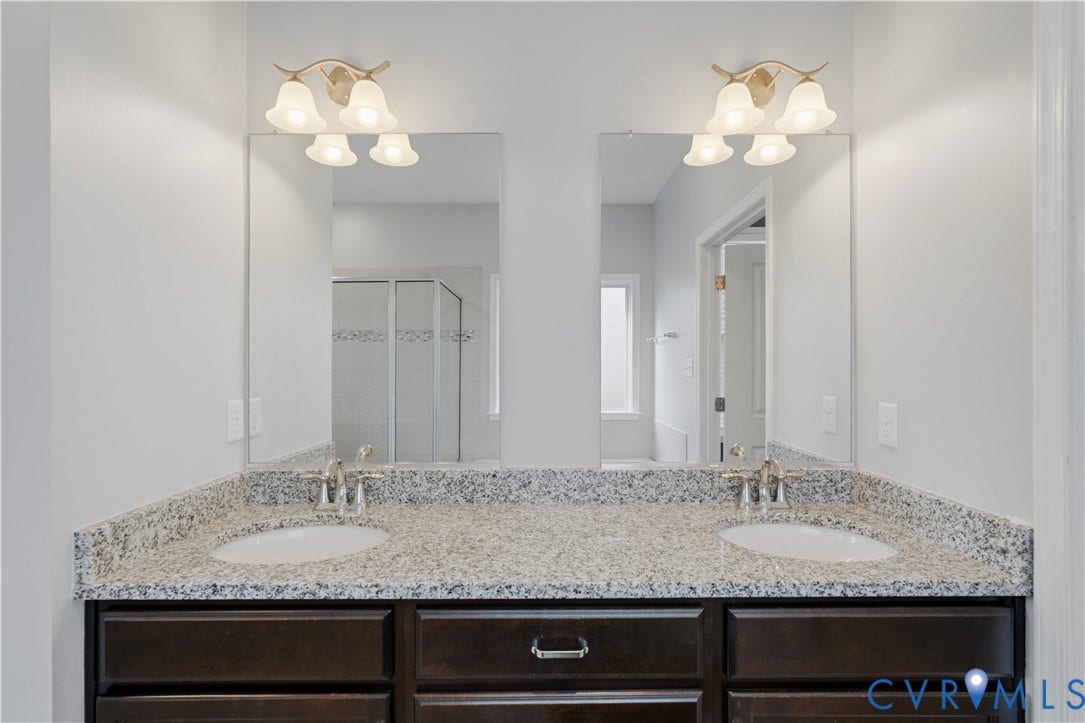 11936 Helmway Court, Chester, VA 23839 Downstairs Primary bathroom. Dual-sink vanity featuring stone-finish countertops, dark wood cabinetry, two separate plate-glass mirrors, and two three-light brushed-nickel fixtures with frosted glass shades