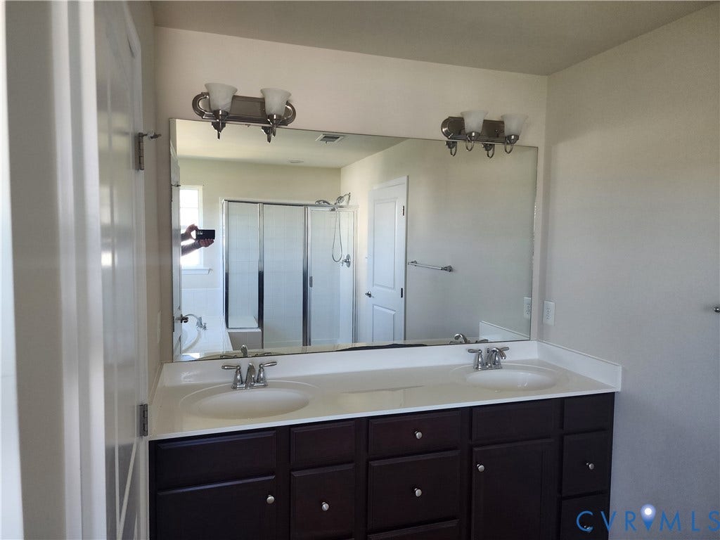 9031 Annex Lane, Mechanicsville, VA 23116 Double vanity with a solid-surface countertop, dual integrated sinks, dark wood-finish cabinetry, a full-width vanity mirror, and dual wall-mounted light fixtures