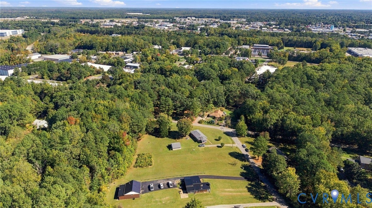 Aerial perspective of an expansive lot featuring mature woodland, open grassy acreage, a paved driveway, and multiple detached structures