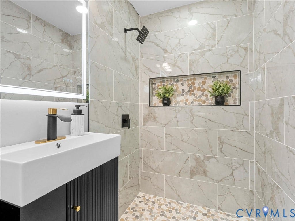 1811 Murdoch Road, Henrico, VA 23229 Bathroom vanity featuring a fluted dark cabinet, white rectangular basin, and matte black faucet, accompanied by a shower enclosure with large-format stone-finish tile walls, pebble-stone flooring, and a recessed tiled niche