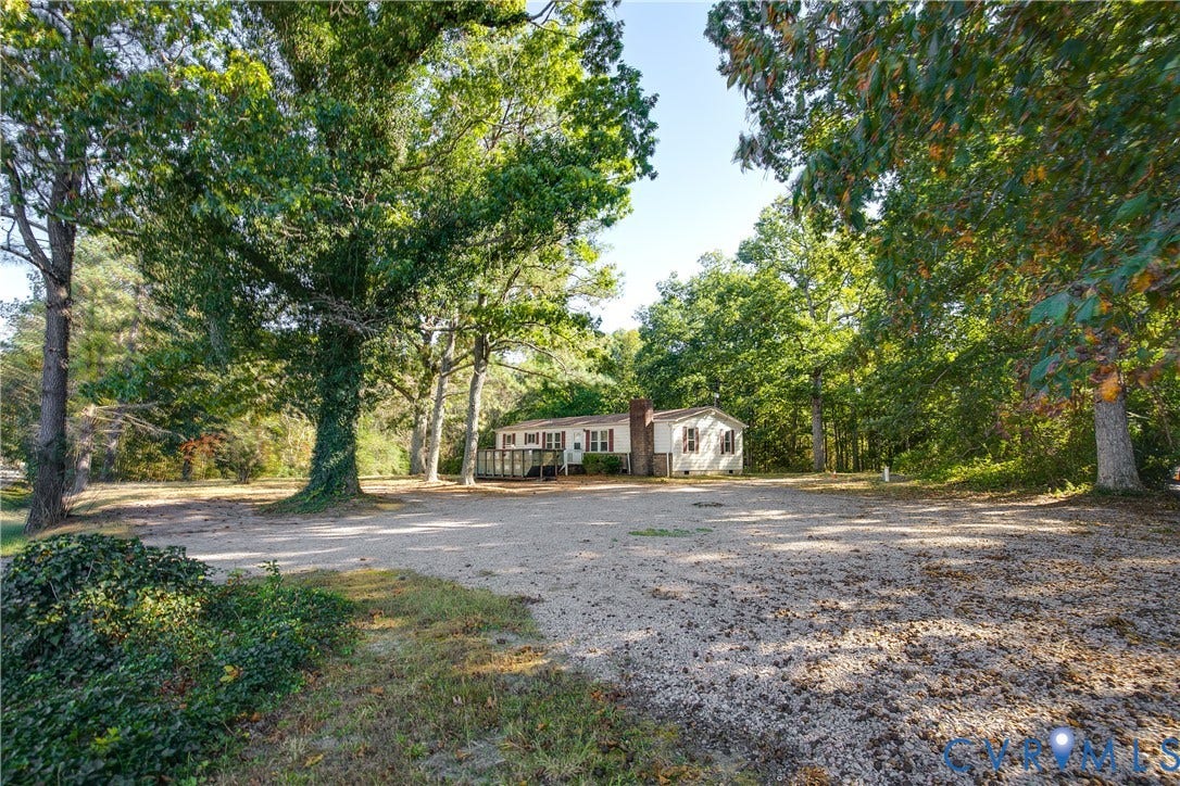 20311 McKenney Highway, Dinwiddie, VA 23882 Single-story exterior featuring light-toned siding, a brick chimney, an elevated wooden deck, and an expansive gravel driveway situated within a mature wooded lot