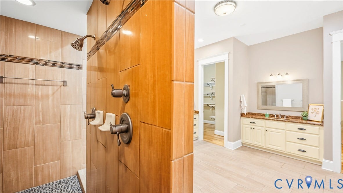 3056 Greywalls Drive, Powhatan, VA 23139 Walk-in shower featuring wood-finish tile walls, stone mosaic floor inlay, and bronze-toned hardware, paired with a dual-sink vanity topped with stone-finish surfacing