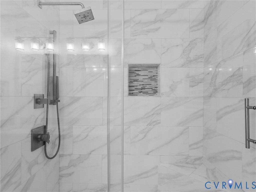 Walk-in shower featuring floor-to-ceiling veined tile, a glass enclosure, a rainfall showerhead with a handheld sprayer, and a recessed tiled niche