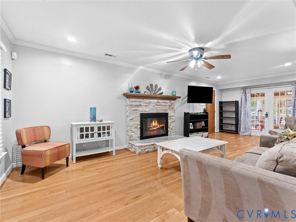 Living area featuring wood-finish flooring, a stacked-stone fireplace with a wooden mantel, a ceiling fan with integrated lighting, and glass-paneled double doors