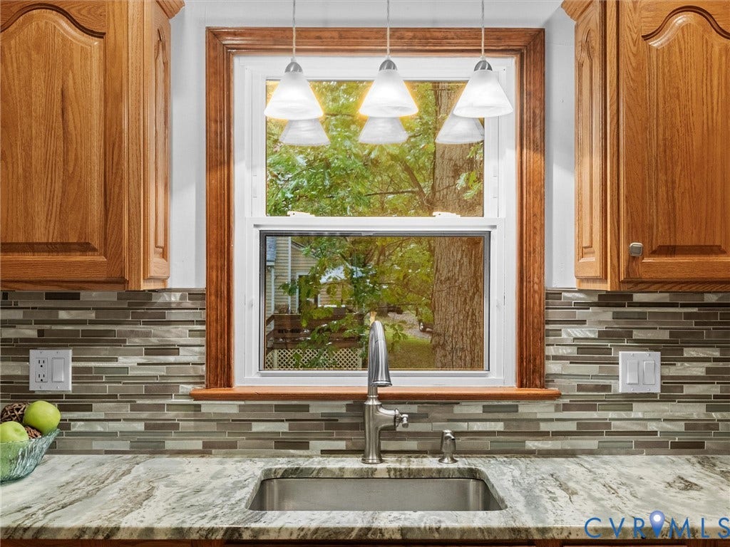 Kitchen features include wood-finish cabinetry, a natural stone countertop, an undermount stainless steel sink with a high-arch faucet, a multi-tonal linear tile backsplash, and triple pendant lighting