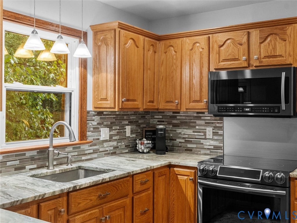Kitchen featuring wood cabinetry, natural stone countertops, multi-tonal glass tile backsplash, stainless steel sink with high-arc faucet, and a black over-the-range microwave