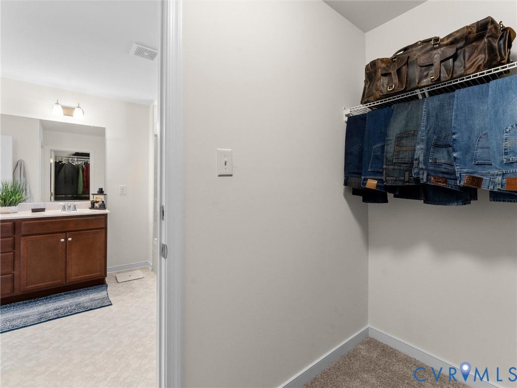 5726 Gossamer Place, Wynwood At FoxCreek, Moseley, VA 23120 Walk-in closet featuring wire shelving, wall-to-wall carpeting, and an entryway into a bathroom equipped with a wood-finish vanity, solid-surface countertop, and wall-mounted vanity lighting