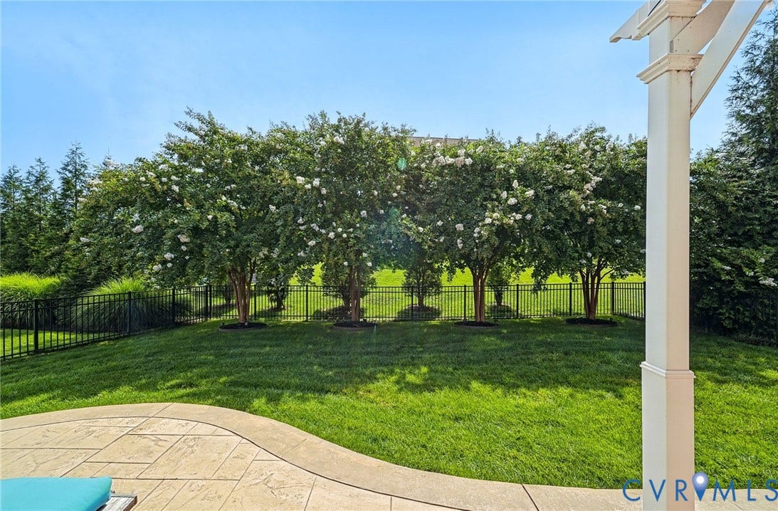 Stamped concrete patio featuring a white pergola, manicured lawn, metal perimeter fencing, and mature flowering trees