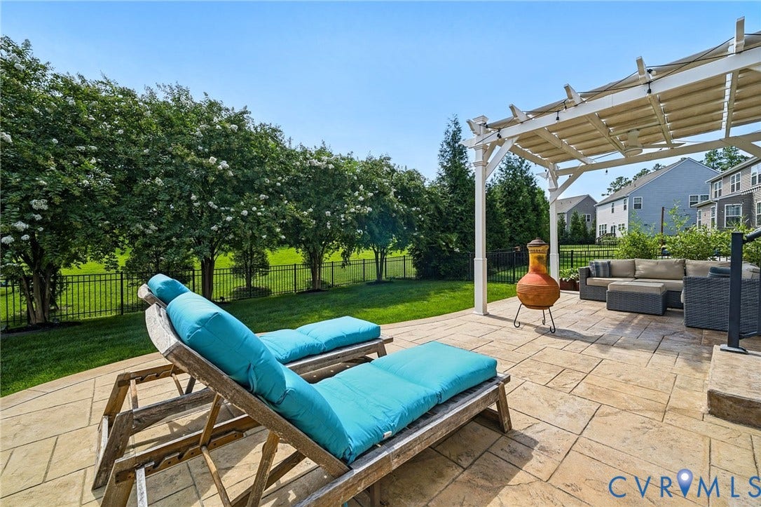 Backyard featuring a paved stone patio, white-painted wood pergola, black metal fencing, and established perimeter trees