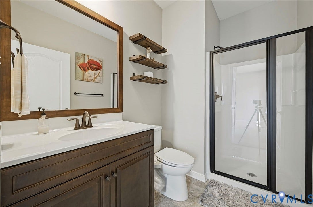 Dark wood cabinetry vanity featuring an integrated basin and countertop, framed wood mirror, wall-mounted wood shelving, and a glass-enclosed shower with a matte black frame