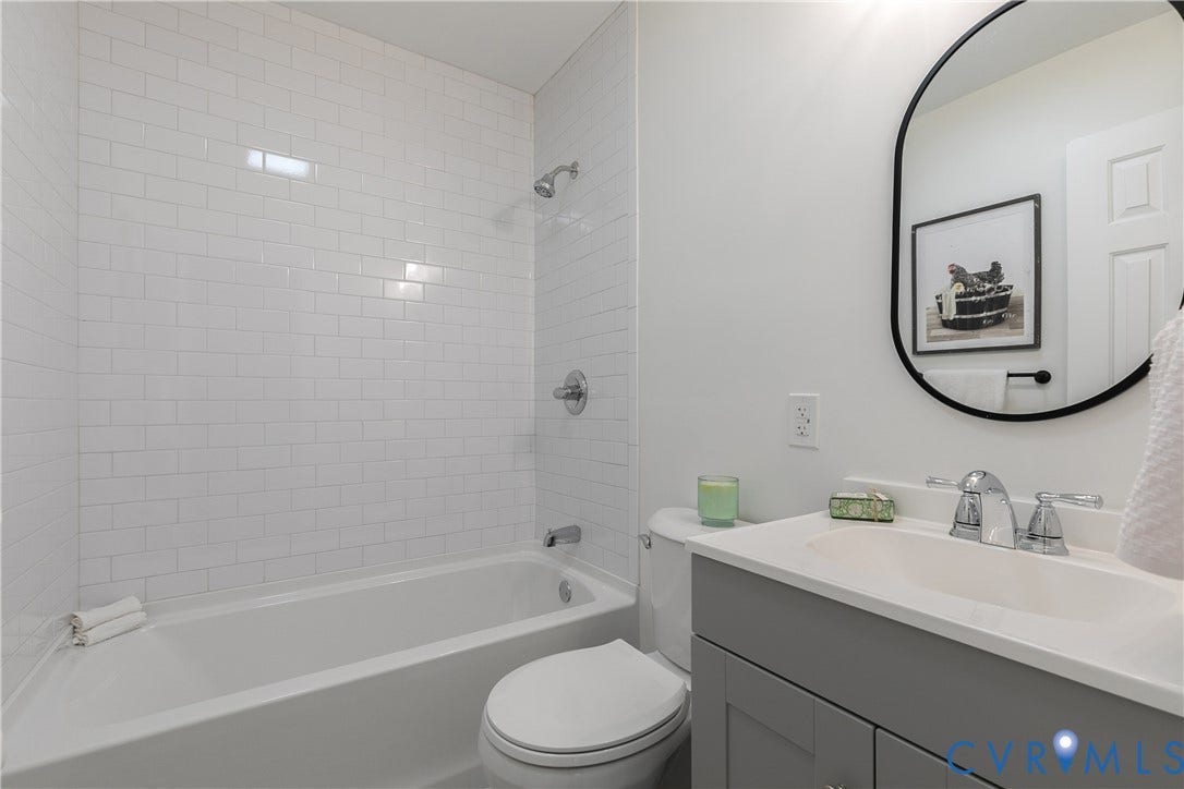 1809 Sussex Street, Whitcomb, Richmond, VA 23223 Bathroom featuring white subway tile tub surround, grey shaker-style vanity, solid surface countertop with integrated sink, chrome hardware, and an oval mirror with a black frame