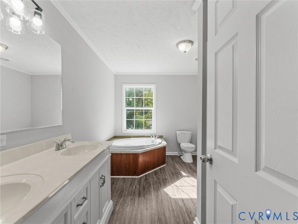 1625 Cedar Lane, Powhatan, VA 23139 Dual-sink vanity with a solid-surface countertop, jetted soaking tub with wood-finish surround, wood-finish flooring, and multi-light wall fixtures