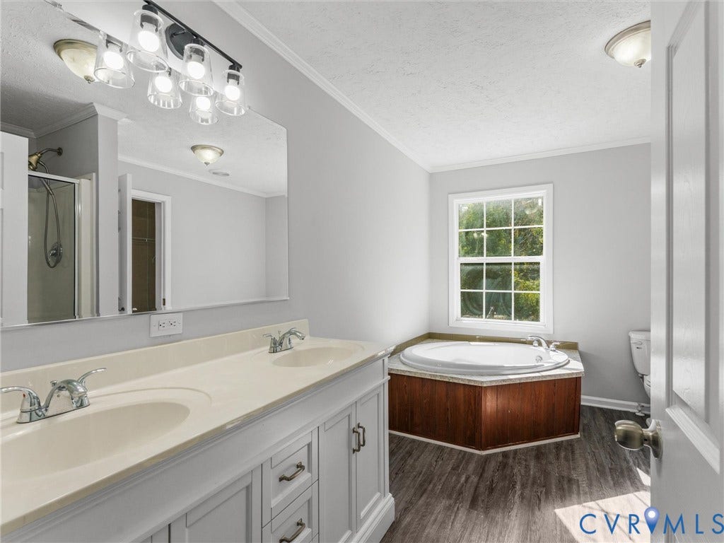 1625 Cedar Lane, Powhatan, VA 23139 Dual-sink vanity with light-toned countertop, large wall-mounted mirror, multi-light fixture, circular jetted soaking tub with wood-finish surround, and wood-finish flooring