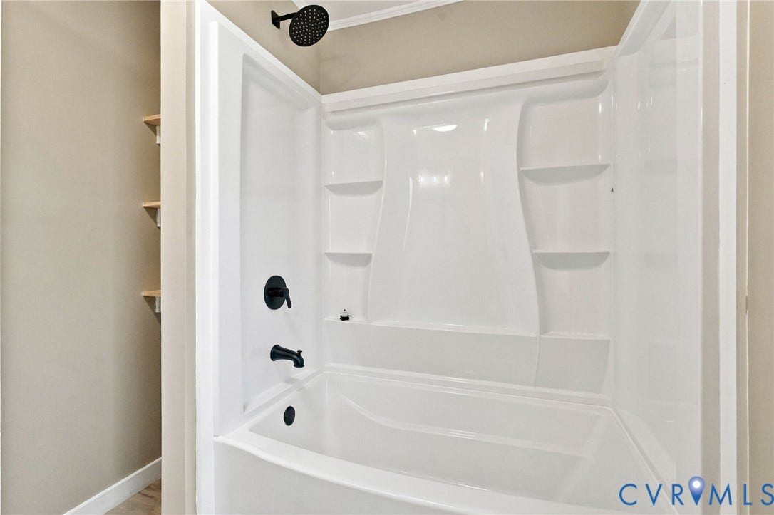 Tub and shower combination featuring a white acrylic surround with integrated shelving, matte black wall-mounted faucet, overhead rainfall showerhead, and wall-mounted wooden shelving units