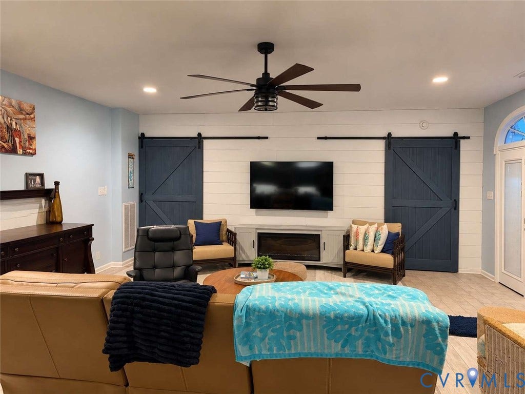 5424 Bellswood Drive, Mechanicsville, VA 23111 Horizontal shiplap accent wall featuring dual sliding barn doors, integrated electric fireplace, matte black ceiling fan, and light-tone tile flooring