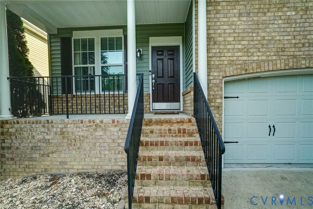 8909 Hollycroft Court, Mechanicsville, VA 23116 Covered front porch featuring a brick facade, decorative iron railings, and a multi-paneled entry door with a metallic kickplate, complemented by a single-car garage with a decorative exterior door