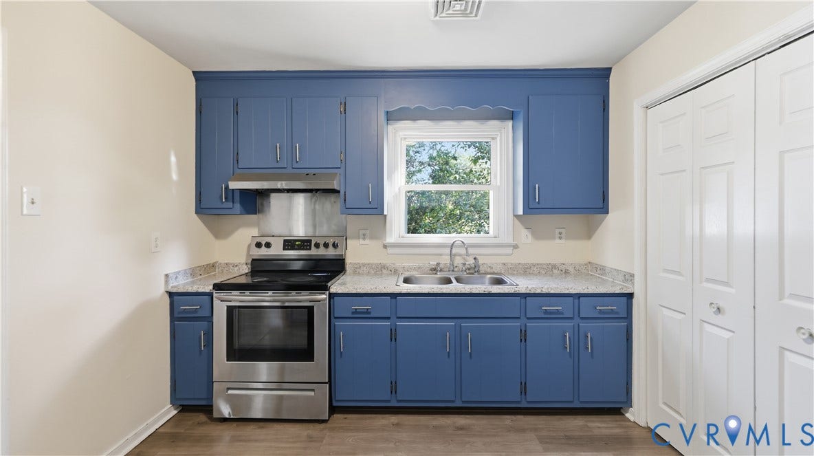 Kitchen featuring blue cabinetry, light-toned stone-finish countertops, stainless steel range with overhead vent, double-basin sink with chrome faucet, and wood-finish flooring