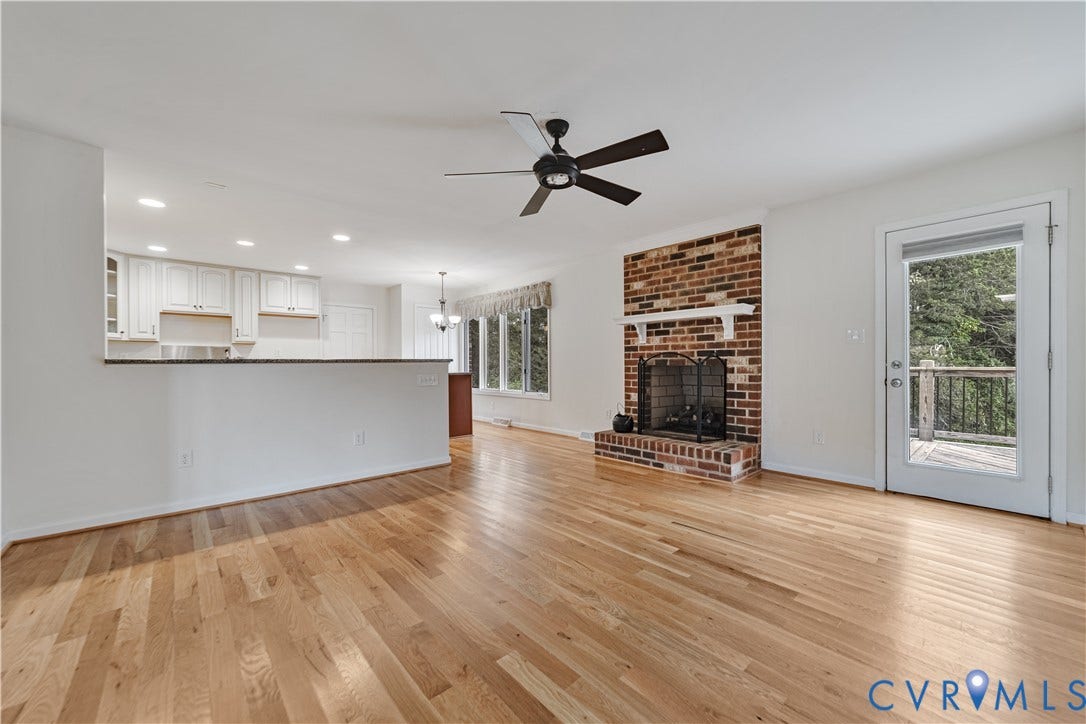Open-concept interior featuring wood-finish flooring, a floor-to-ceiling brick fireplace with a mantel, white kitchen cabinetry, recessed lighting, and a glass-paneled exterior door