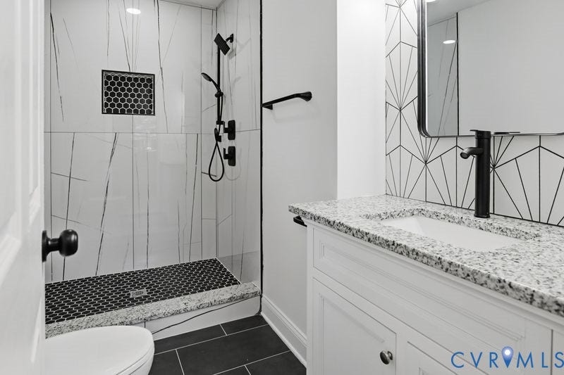 Bathroom featuring a walk-in shower with large-format tile, a stone-finish countertop with an integrated sink, matte black plumbing fixtures, and geometric tile backsplashes