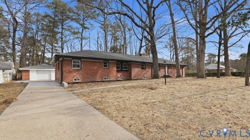 Red brick exterior featuring a hip roof, concrete driveway, detached single-car garage, and mature trees on the lot