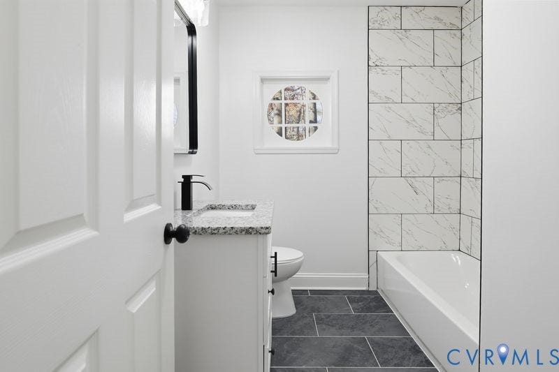 Bathroom featuring a vanity with a stone-finish countertop, matte black faucet, rectangular wall-mounted mirror, tub surround with large-format patterned tile, and dark floor tile