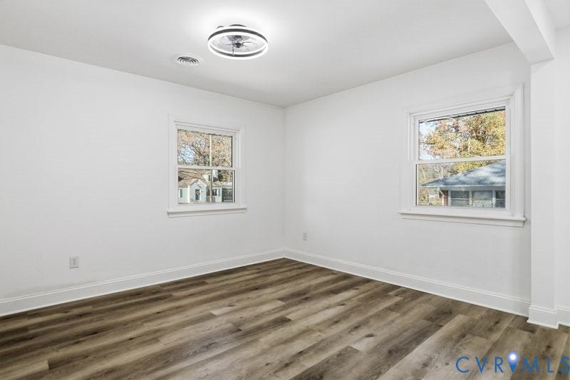 Neutral interior space featuring wood-finish flooring, white walls, dual windows with white casing, and a modern circular flush-mount ceiling fan
