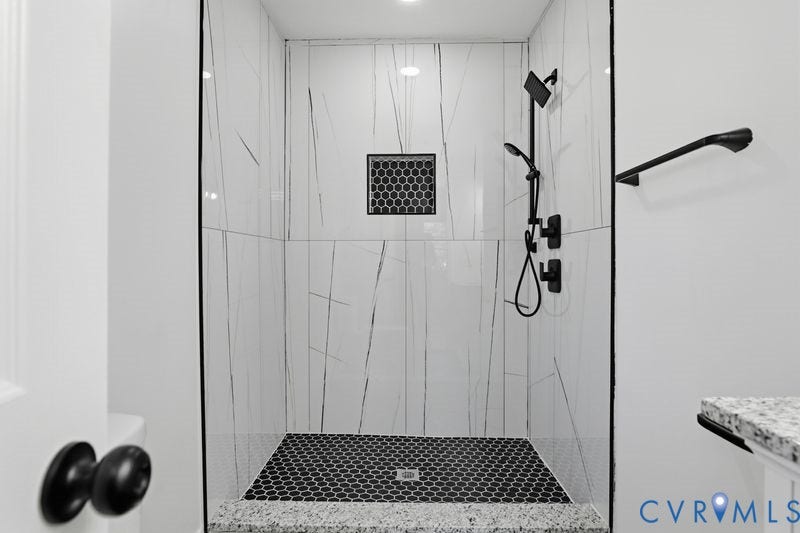 Walk-in shower featuring white large-format wall tile with contrasting veining, matte black fixtures, integrated hexagonal tile niche, and a contrasting dark hexagonal floor tile