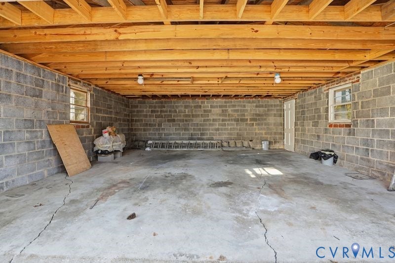 Unfinished utility space featuring exposed wood ceiling joists, concrete block masonry walls, concrete flooring, and two windows