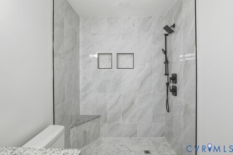 Walk-in shower featuring floor-to-ceiling stone-patterned tile, matte black rainfall shower head with handheld attachment, built-in corner bench, and dual recessed wall niches