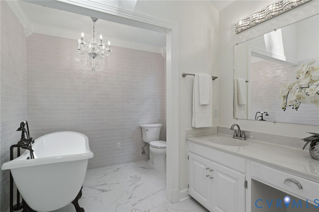 2311 Castle Hill Road, Midlothian, VA 23113 Freestanding soaking tub with a floor-mounted faucet, crystal chandelier, wall-to-wall light-toned subway tile, stone-patterned tile flooring, and a white vanity with integrated lighting