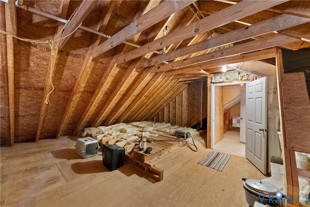 Unfinished attic space featuring exposed wooden structural rafters, plywood subflooring, and a six-panel interior door