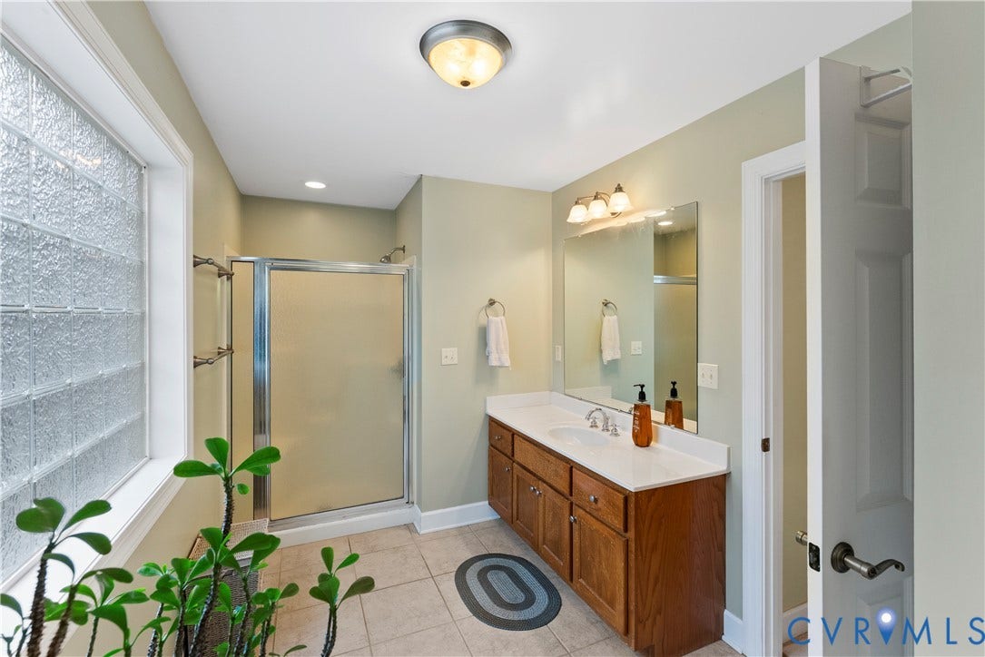 Bathroom featuring a glass block window, stall shower with a metal frame, wood-finish vanity cabinet, solid-surface countertop with an integrated sink, and large wall-mounted mirror with attached lighting