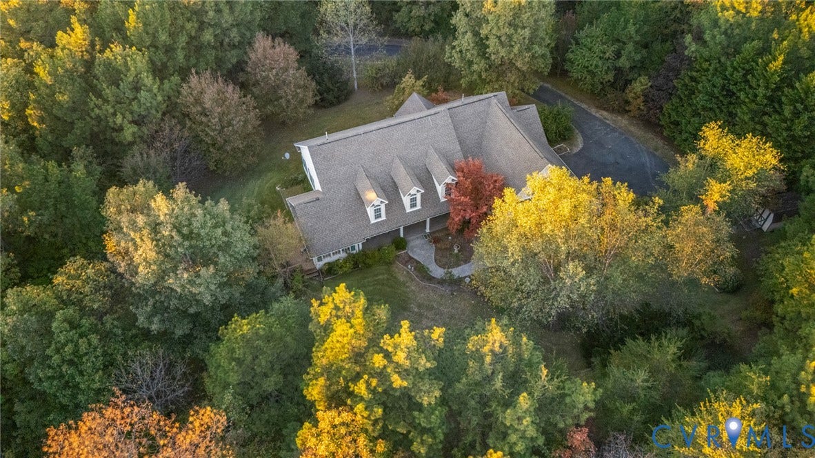 Aerial perspective of a multi-gabled residence featuring a paved driveway, shingled roofing, three dormer windows, and a paved walkway, all situated on a heavily wooded lot
