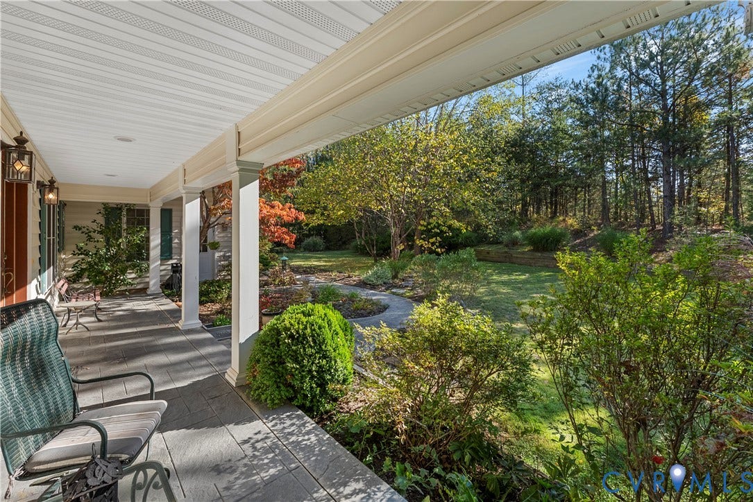 Covered porch with stone-style tile flooring, decorative wall-mounted lighting, and column supports, overlooking a landscaped backyard featuring a stone-paver walkway and mature trees