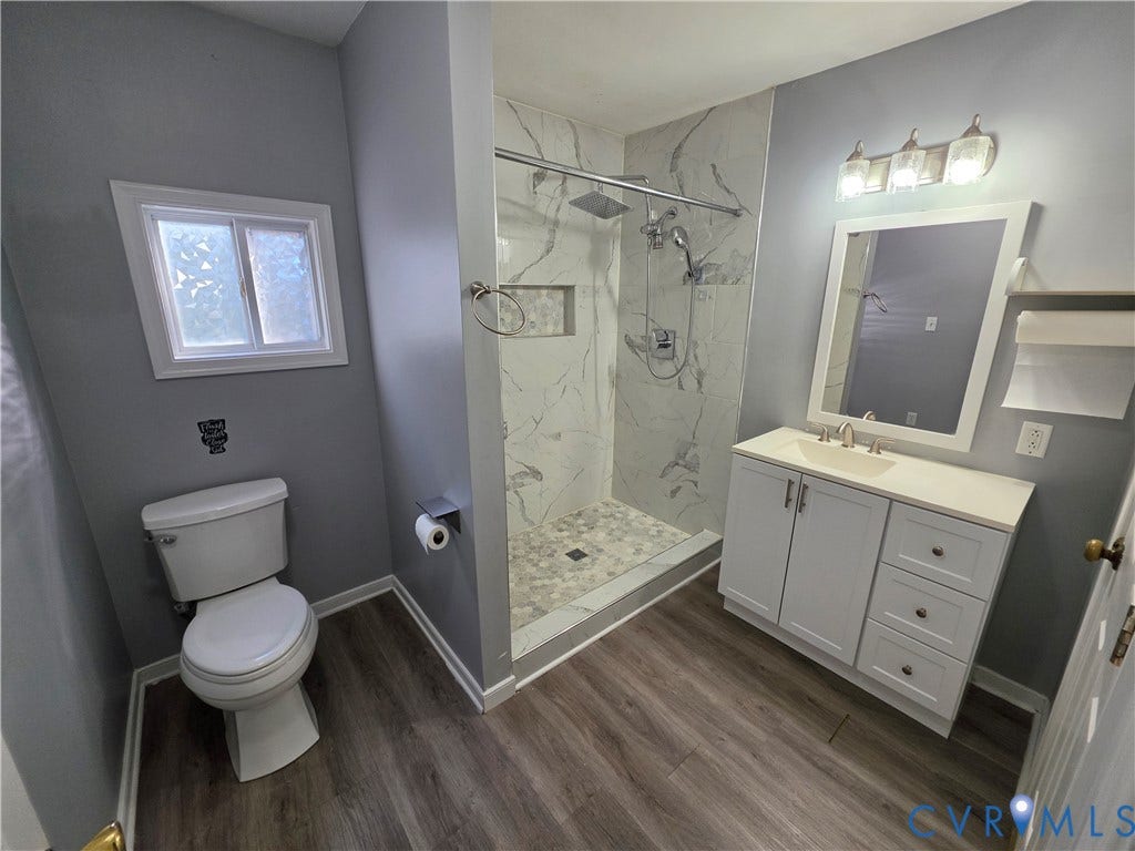 2406 Trenton Street, Hopewell, VA 23860 Bathroom featuring a shower stall with stone-patterned tile, built-in shelving niche, overhead rain shower head, wood-finish flooring, and white vanity cabinetry with a solid surface countertop
