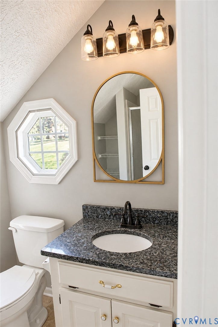 4229 Cedar Creek Lane, Prince George, VA 23875 Vanity featuring a stone-composite countertop with an integrated basin, arched gold-framed mirror, four-light glass fixture, and an octagonal window
