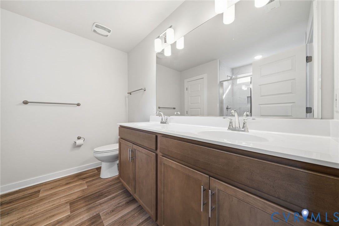 8209 Grantham Woods Way, Grantham Woods, Henrico, VA 23231 Dual-sink vanity featuring a white countertop, wood-finish cabinetry, full-width mirror, and multi-bulb light fixture, accompanied by wood-finish flooring