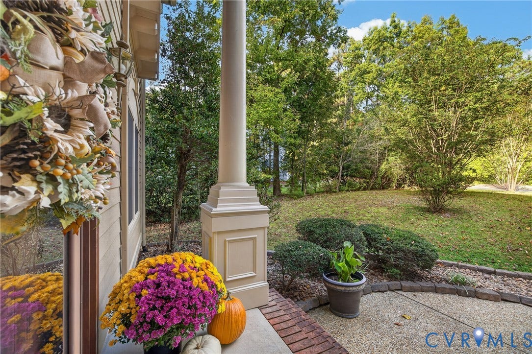 Covered porch featuring a paneled structural column, aggregate concrete flooring, and a brick border, opening to a grass-covered backyard with mature trees