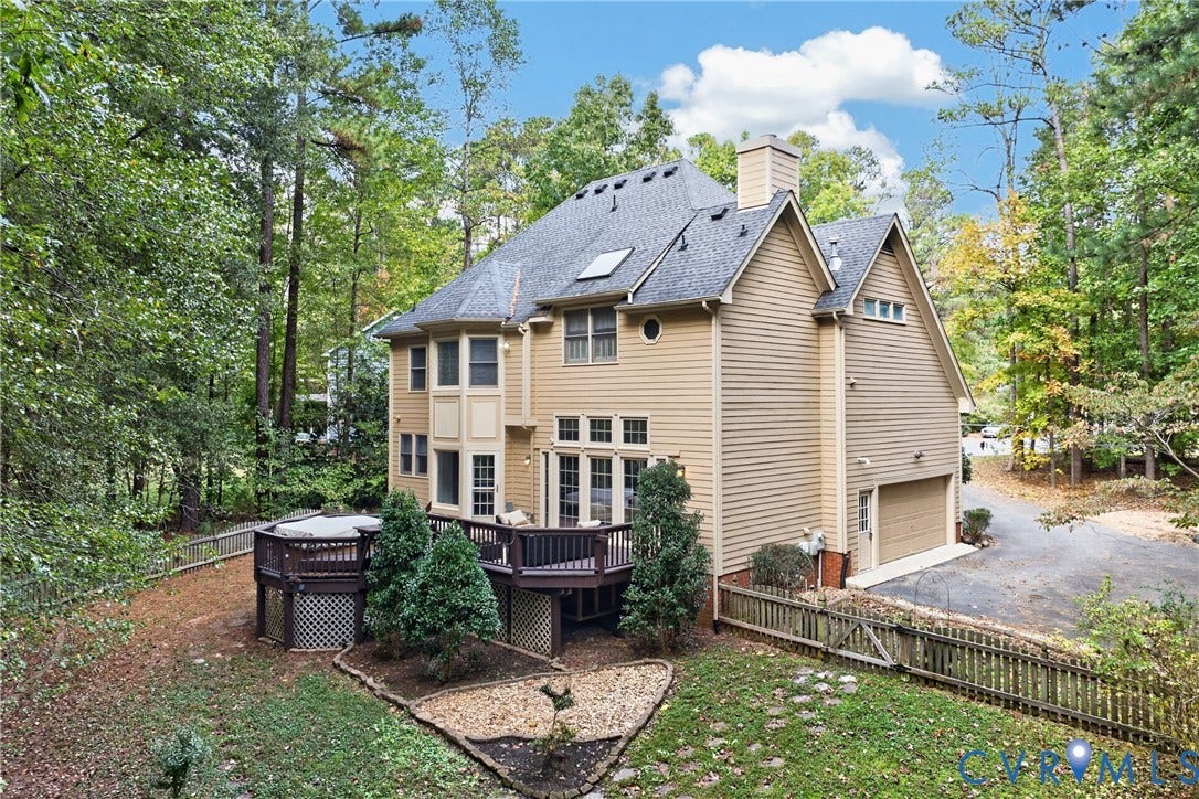 Multi-story residence featuring light-toned horizontal siding, an elevated wood deck, a roof-integrated skylight, and an attached side-entry garage, set on a wooded lot with a perimeter fence