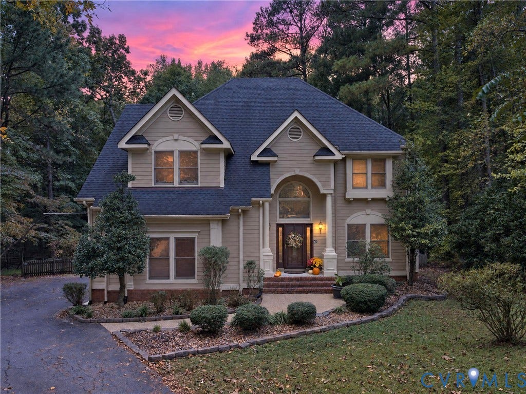 Two-story exterior featuring tan horizontal siding, an arched entryway with column accents, a multi-gabled roofline, and landscaped brick-bordered garden beds