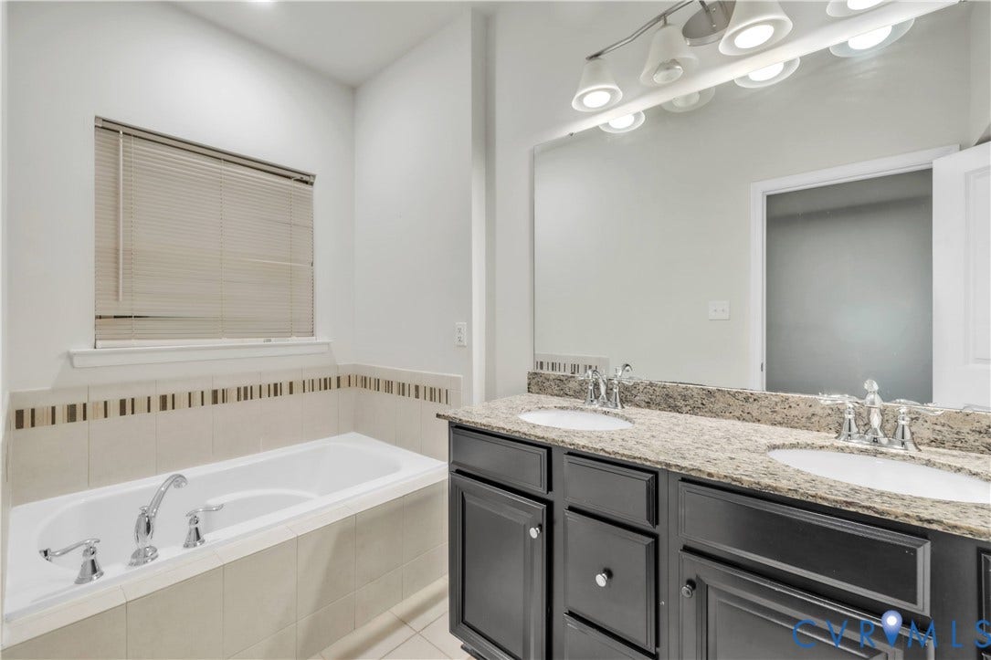 4511 Wistar Woods Trail, Richmond, VA 23228 Dual-sink vanity with dark cabinetry, stone-finish countertop, soaking tub with tile surround, and expansive wall-mounted vanity mirror
